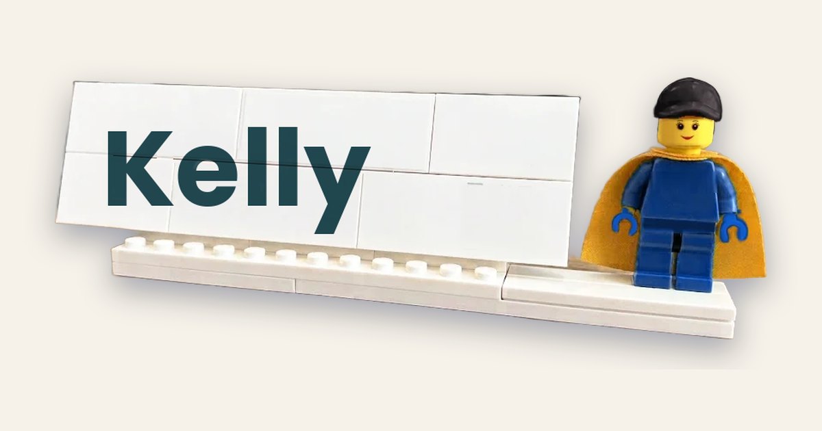 A white Lego name plaque printed with the name Kelly, with a minifig in a yellow cape standing beside it